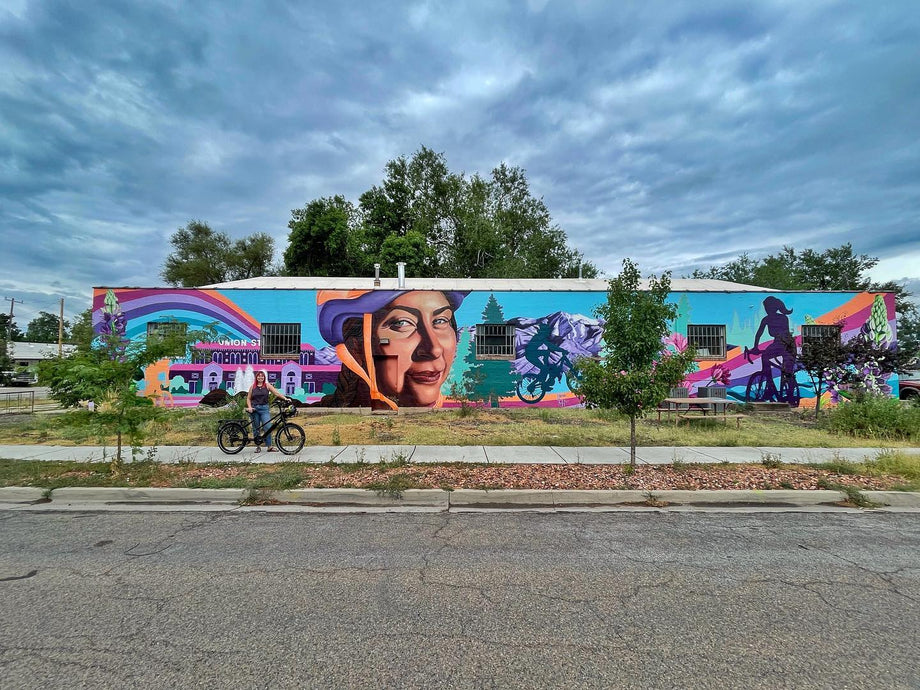 Artist, Lindsay Huss, stands in front of the mural she painted on the Ogden Bicycle Collective building in 2022