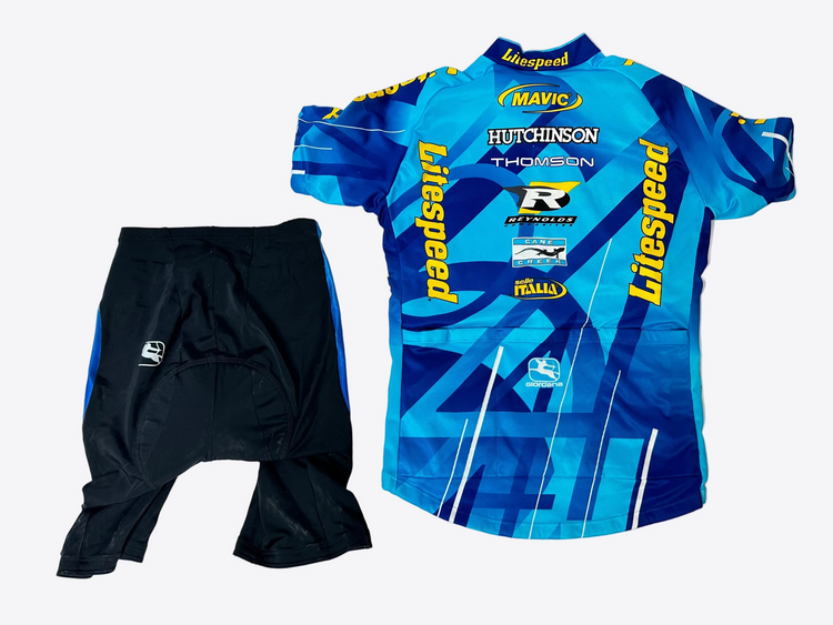 Blue cycling jersey with various brand logos and black shorts on a light gray background