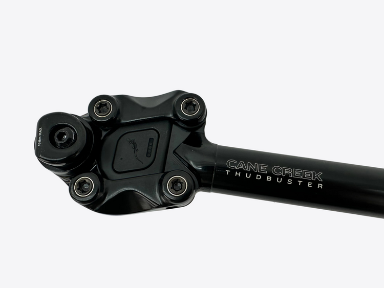A black Cane Creek Thudbuster bicycle headset component with visible ball bearings and silver branding text, photographed on a light gray tiled surface.