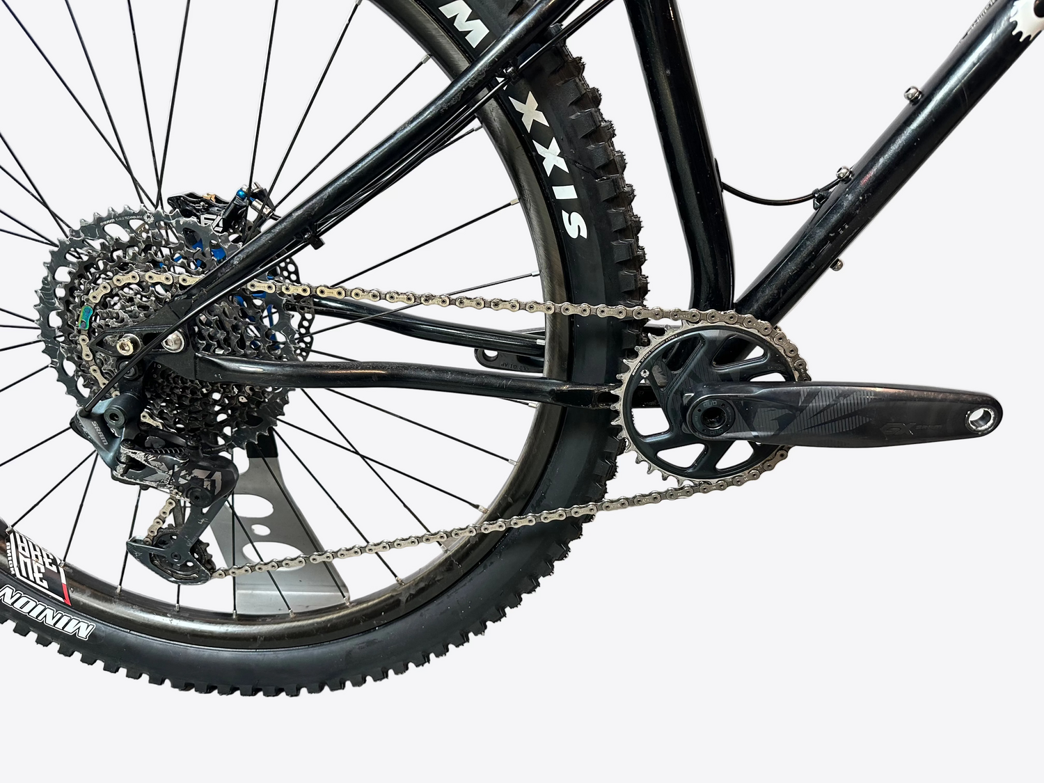 Close-up of a black mountain bike's rear wheel, chainring, chain, and cassette, photographed indoors against a light gray backdrop.