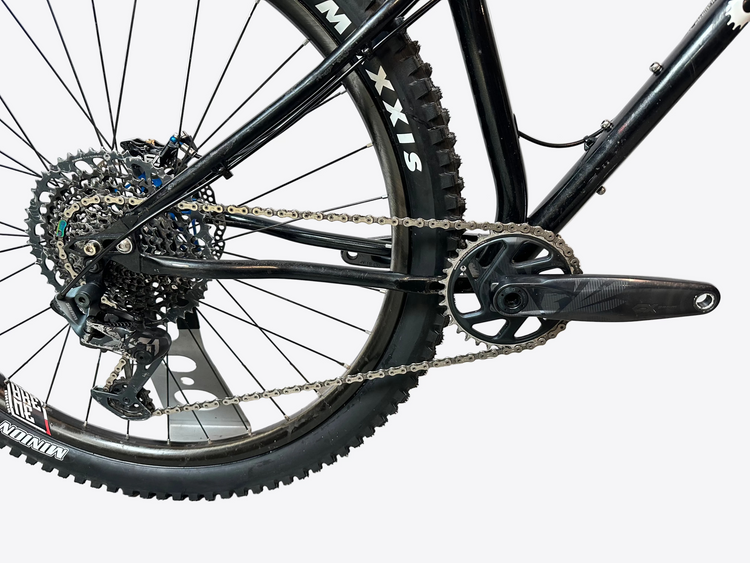 Close-up of a black mountain bike's rear wheel, chainring, chain, and cassette, photographed indoors against a light gray backdrop.