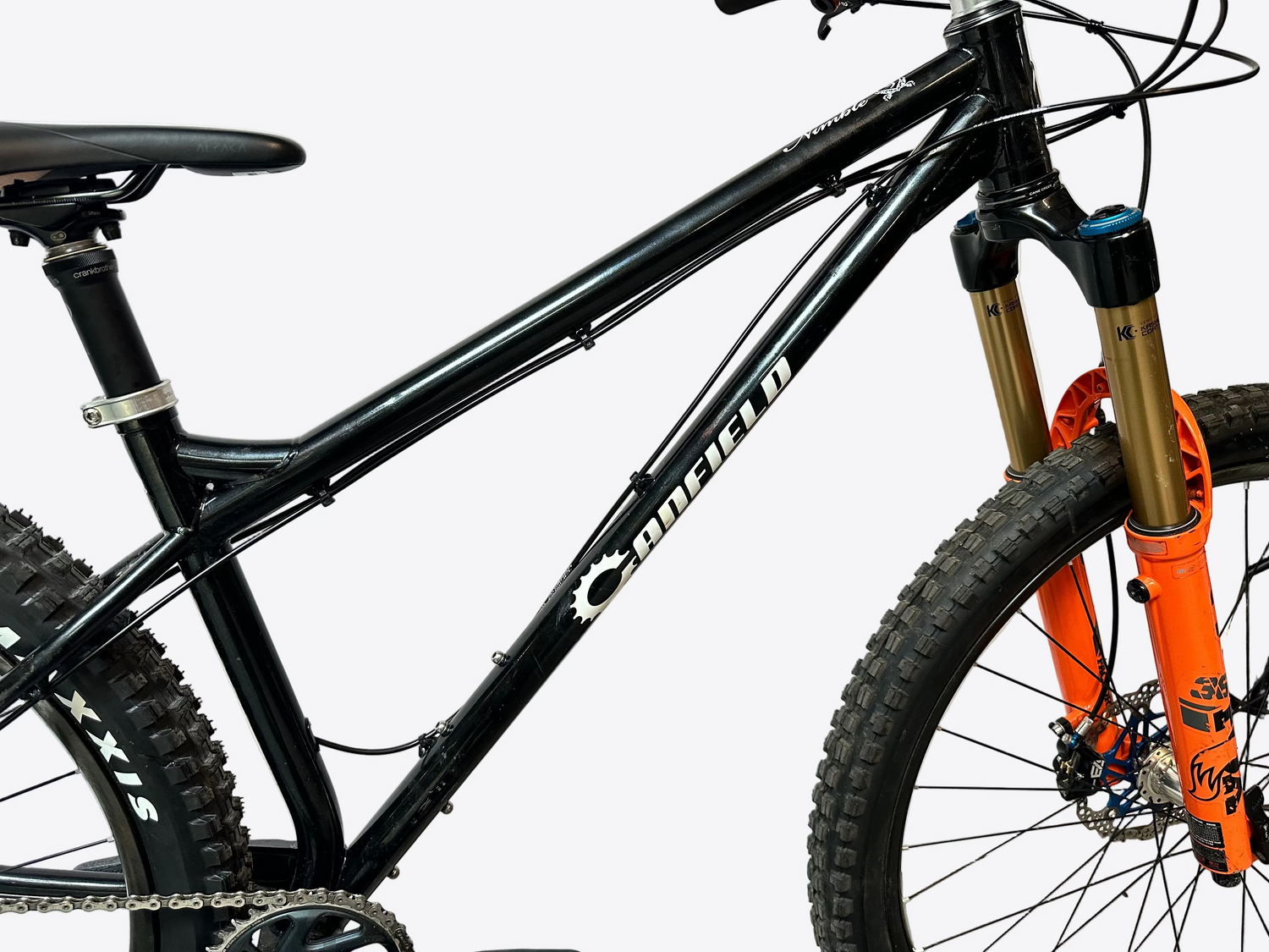 Close-up of a black mountain bike frame with a white Sunfield logo, gold and orange front suspension fork, and knobby tires against a plain light background.