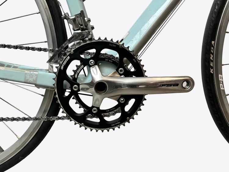 Close-up of a bicycle chainring and chain on a light blue bike with a white wall background.