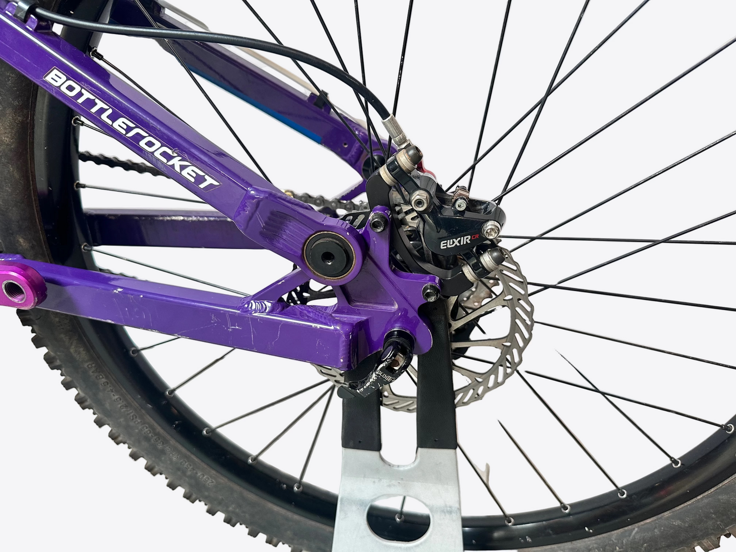 Close-up of a purple bicycle frame with 'Bottle Rocket' branding, focusing on the wheel and chain mechanism.