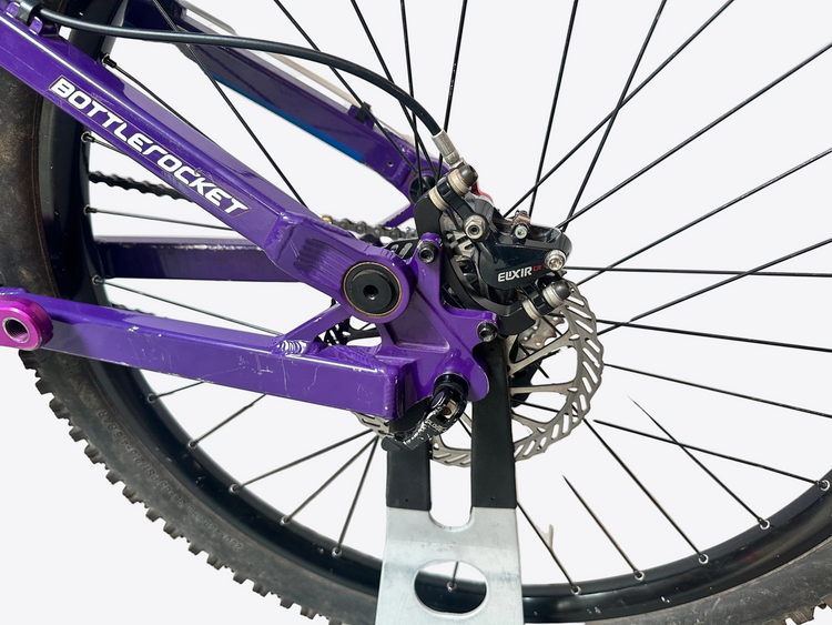 Close-up of a purple bicycle frame with 'Bottle Rocket' branding, focusing on the wheel and chain mechanism.