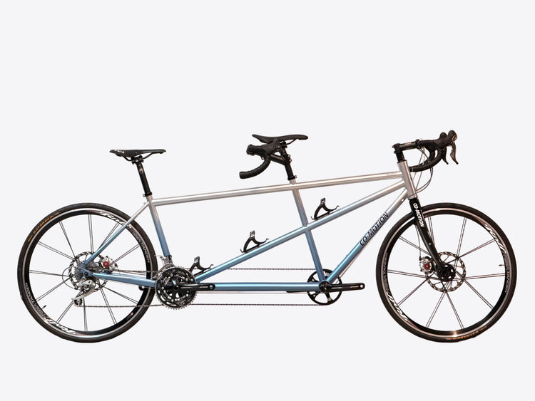 A long tandem bicycle with a silver and light blue frame, black drop handlebars, and black wheels, photographed against a white studio backdrop.