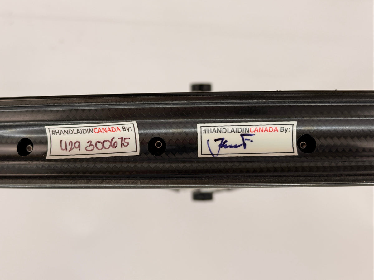 Close-up of a carbon fiber rim showing two white stickers with handwritten and printed text, including 'HANDLAIDIN CANADA BY:' and model markings.