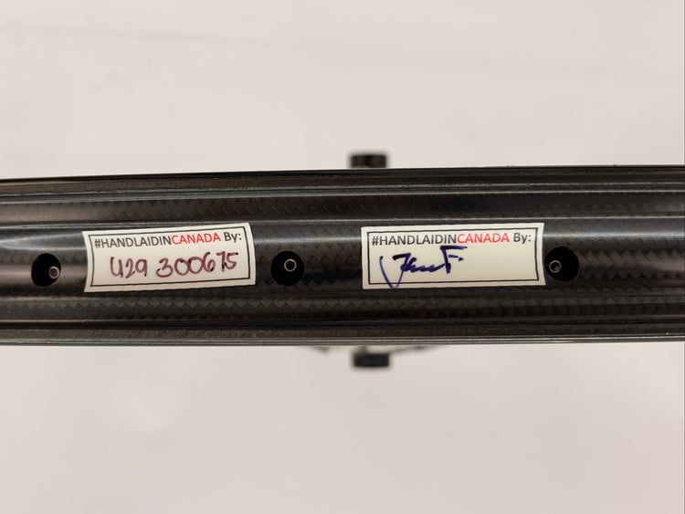 Close-up of a carbon fiber rim showing two white stickers with handwritten and printed text, including 'HANDLAIDIN CANADA BY:' and model markings.